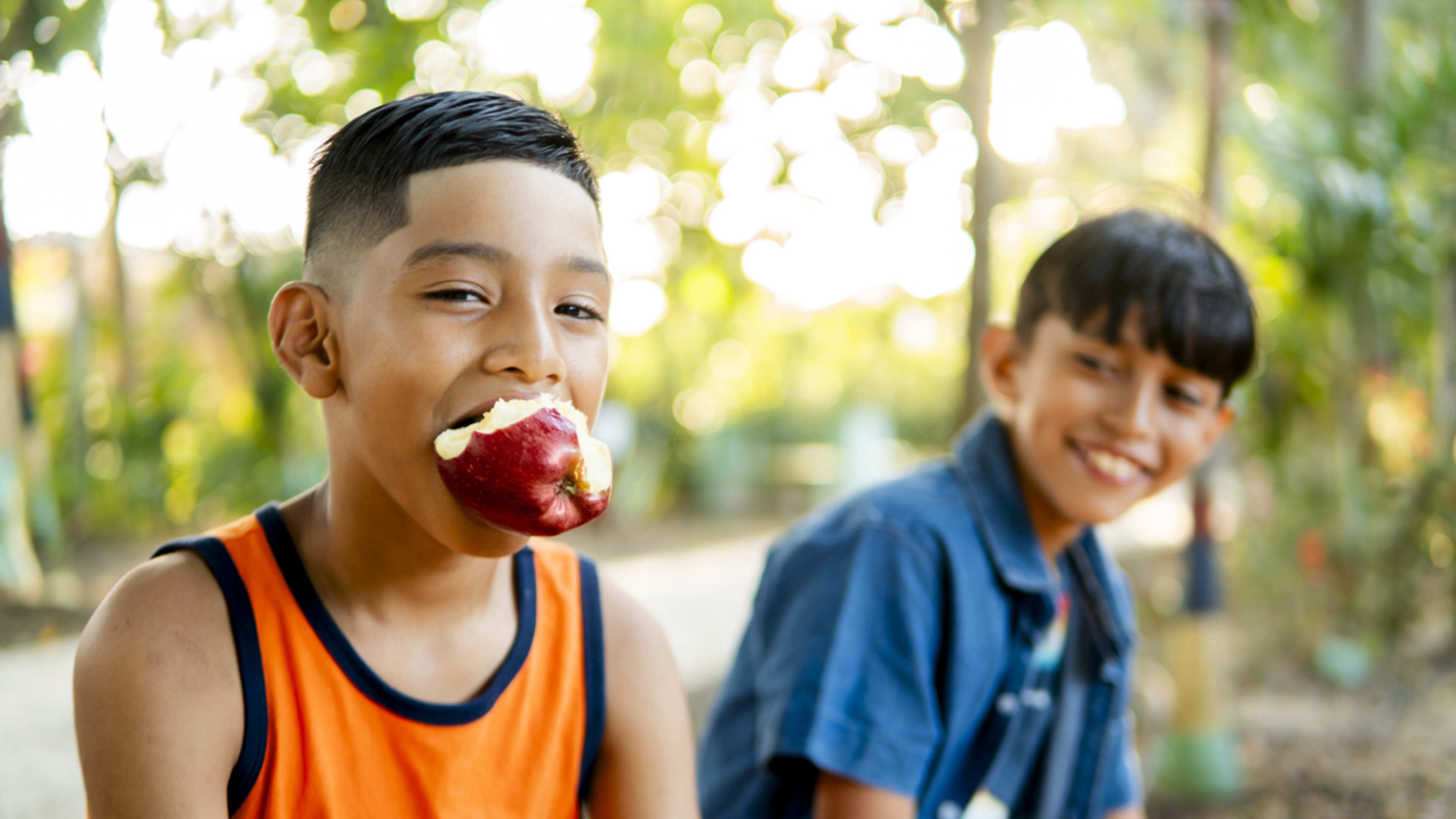 two boys sitting, one has a half eaten apple in his mouth