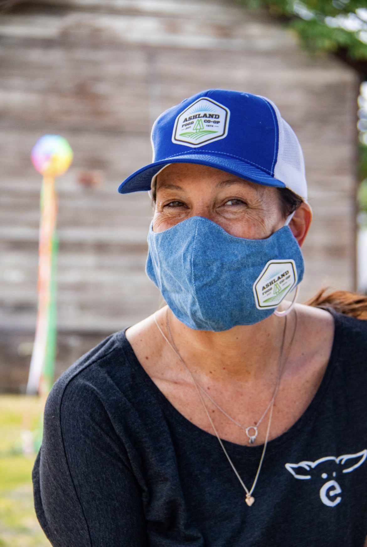 Photo Credit: Chelsea Whitney Art Julie O'Dwyer, Ashland Food Co-op (AFC) Board Vice President, smiles from behind a teal mask with an AFC sticker and AFC branded hat