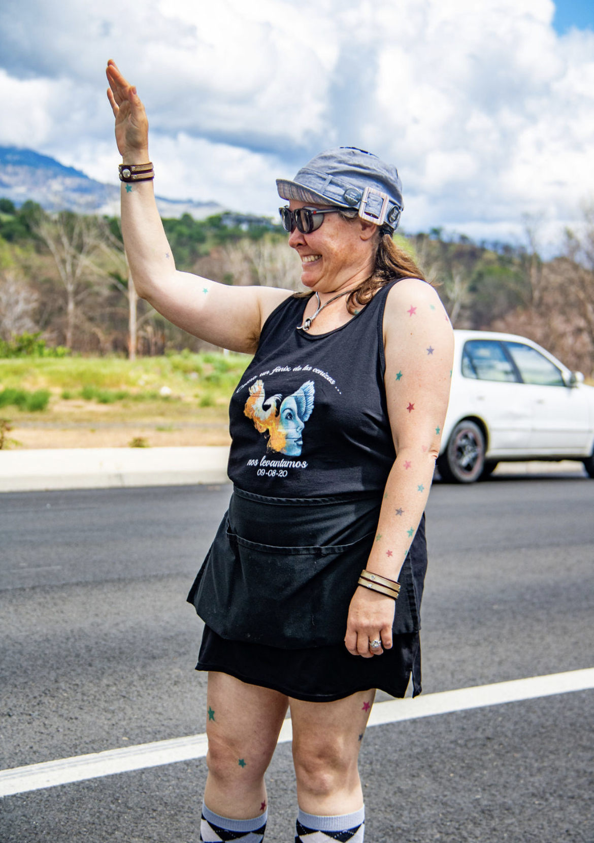 Photo Credit: Chelsea Whitney Art a woman stands grinning and waving to someone in the distance. She has star shaped stickers on her bare arms and legs and is holding a stack of Zone Captain flyers.