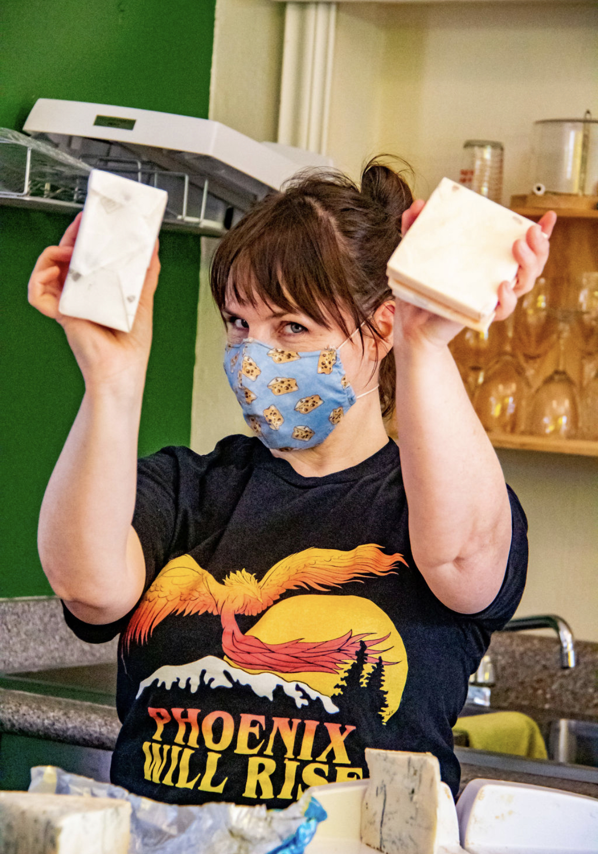Photo Credit: Chelsea Whitney Art a cheese seller smiles from behind her mask as she holds up two blocks of cheese to the camera